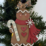 The Rustic Rhode's DIY Victorian Gingerbread Candy Cane Ornament features unassembled pieces for a gingerbread person with a red scarf, two red hearts, and a candy cane, hanging on a festive Christmas tree with green branches and a bow.