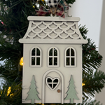 The Rustic Rhode’s DIY Victorian Gingerbread House Ornament kit features a wooden house with a heart-shaped door, two green trees, and patterned roof. Includes plaid ribbon and unassembled pieces—perfect to paint or craft for your Christmas tree!.