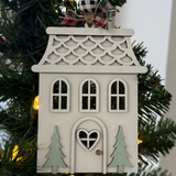 The Rustic Rhode’s DIY Victorian Gingerbread House Ornament kit features a wooden house with a heart-shaped door, two green trees, and patterned roof. Includes plaid ribbon and unassembled pieces—perfect to paint or craft for your Christmas tree!.