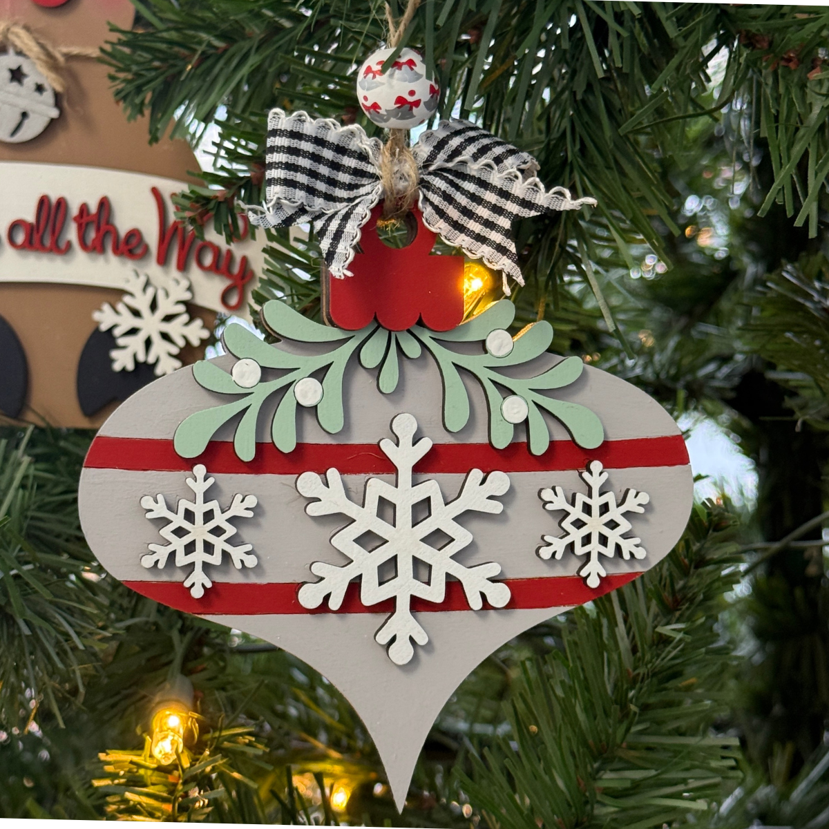The Rustic Rhode's DIY Mistletoe Ornament—Snowflakes features a gray and red design with white snowflakes, green leaves, and a checkered bow, perfect for painting at home or gifting as an unassembled ornament kit. Warm lights add festive cheer.
