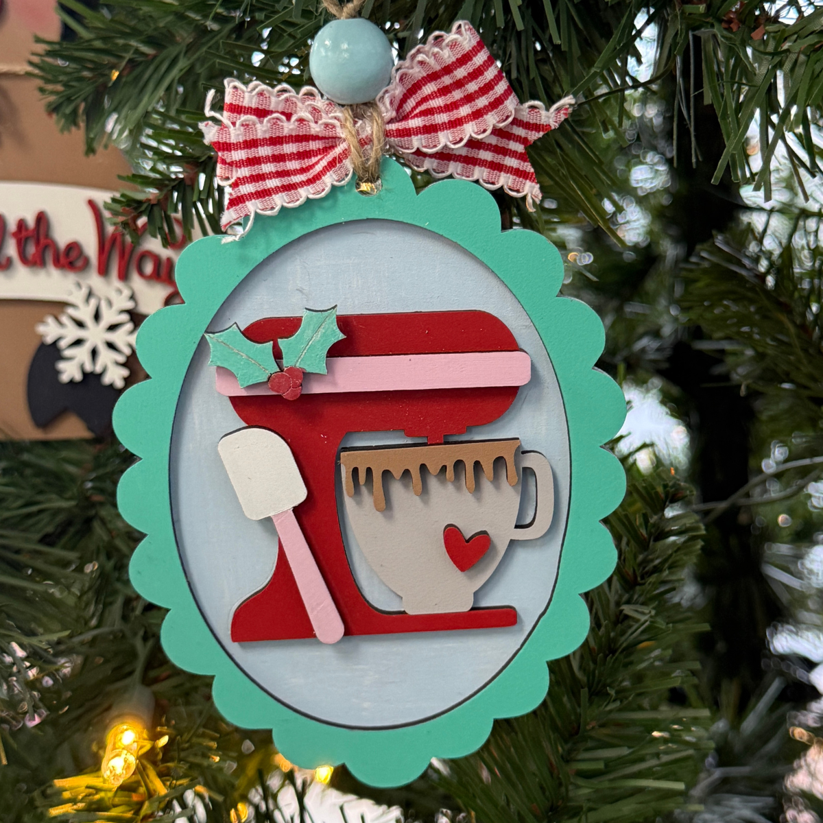 The Rustic Rhode DIY Christmas Baking Mixer Ornament features an oval shape with a red stand mixer, heart mug, white spatula, and holly leaves. Kit includes unassembled pieces and a red & white ribbon for hanging.