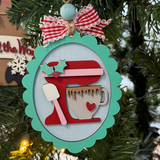 The Rustic Rhode DIY Christmas Baking Mixer Ornament features an oval shape with a red stand mixer, heart mug, white spatula, and holly leaves. Kit includes unassembled pieces and a red & white ribbon for hanging.