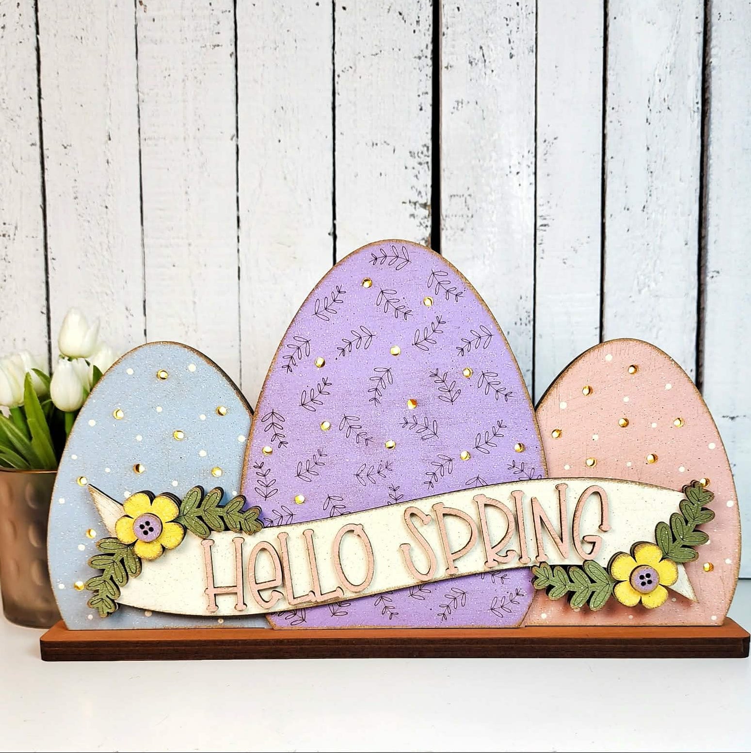 A wooden sign by The Rustic Rhode (P. Fallon Group) features pastel blue, purple, and pink eggs with "HELLO SPRING" text and floral accents—ideal for a seasonal project menu. White tulips in a copper vase sit in the background.