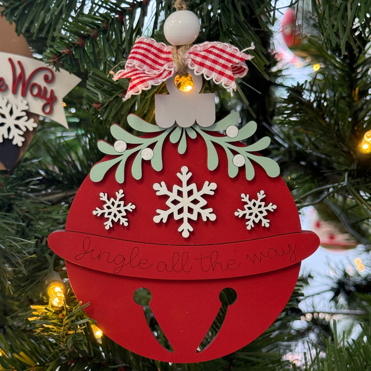 A DIY Mistletoe Ornament-Bell from The Rustic Rhode, featuring red jingle bell style with white snowflakes, green leaves, and a red/white bow, hangs on a Christmas tree—ready to personalize at home using unassembled kit pieces.