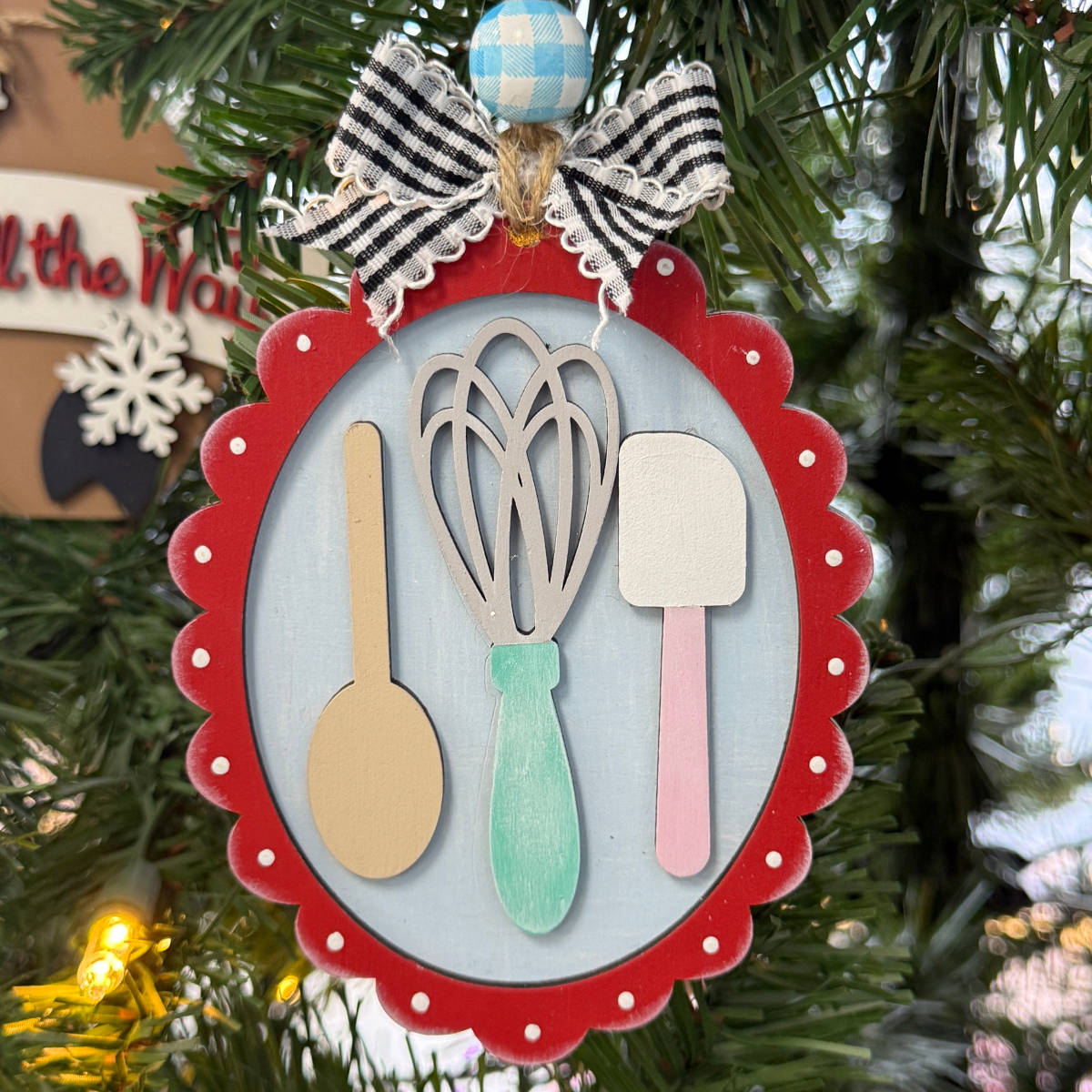 The Rustic Rhode DIY Christmas Baking Utensils Ornament features a red scalloped frame with a wooden spoon, whisk, and spatula, topped with gingham and blue-checkered bows—perfect for painting or decorating your own festive tree.