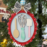 The Rustic Rhode DIY Christmas Baking Utensils Ornament features a red scalloped frame with a wooden spoon, whisk, and spatula, topped with gingham and blue-checkered bows—perfect for painting or decorating your own festive tree.