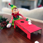 A playful holiday scene features The Rustic Rhode DIY Elf Prop Kit, with a toy elf in festive clothes playing a miniature red cornhole game. Handcrafted props and pine-and-berry holiday decorations complete this charming display.