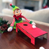 A playful holiday scene features The Rustic Rhode DIY Elf Prop Kit, with a toy elf in festive clothes playing a miniature red cornhole game. Handcrafted props and pine-and-berry holiday decorations complete this charming display.