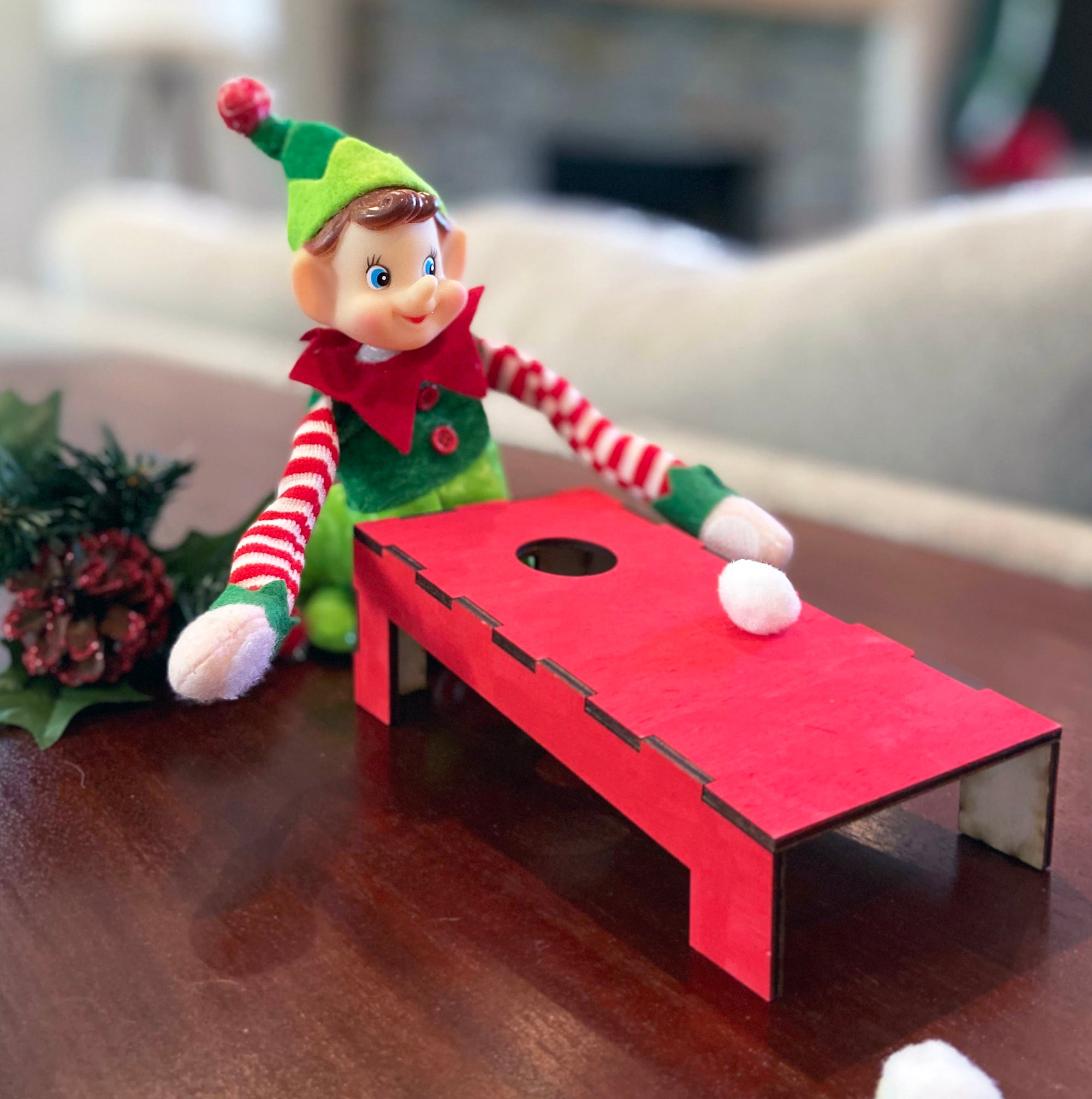 A playful holiday scene features The Rustic Rhode DIY Elf Prop Kit, with a toy elf in festive clothes playing a miniature red cornhole game. Handcrafted props and pine-and-berry holiday decorations complete this charming display.