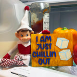 An elf doll from The Rustic Rhode’s DIY Elf Prop Kit sits in a fridge beside veggies, holding an “I AM JUST CHILLIN OUT” sign with ice cubes. A ketchup bottle appears in the background—ideal for festive displays or creative elf scenes.