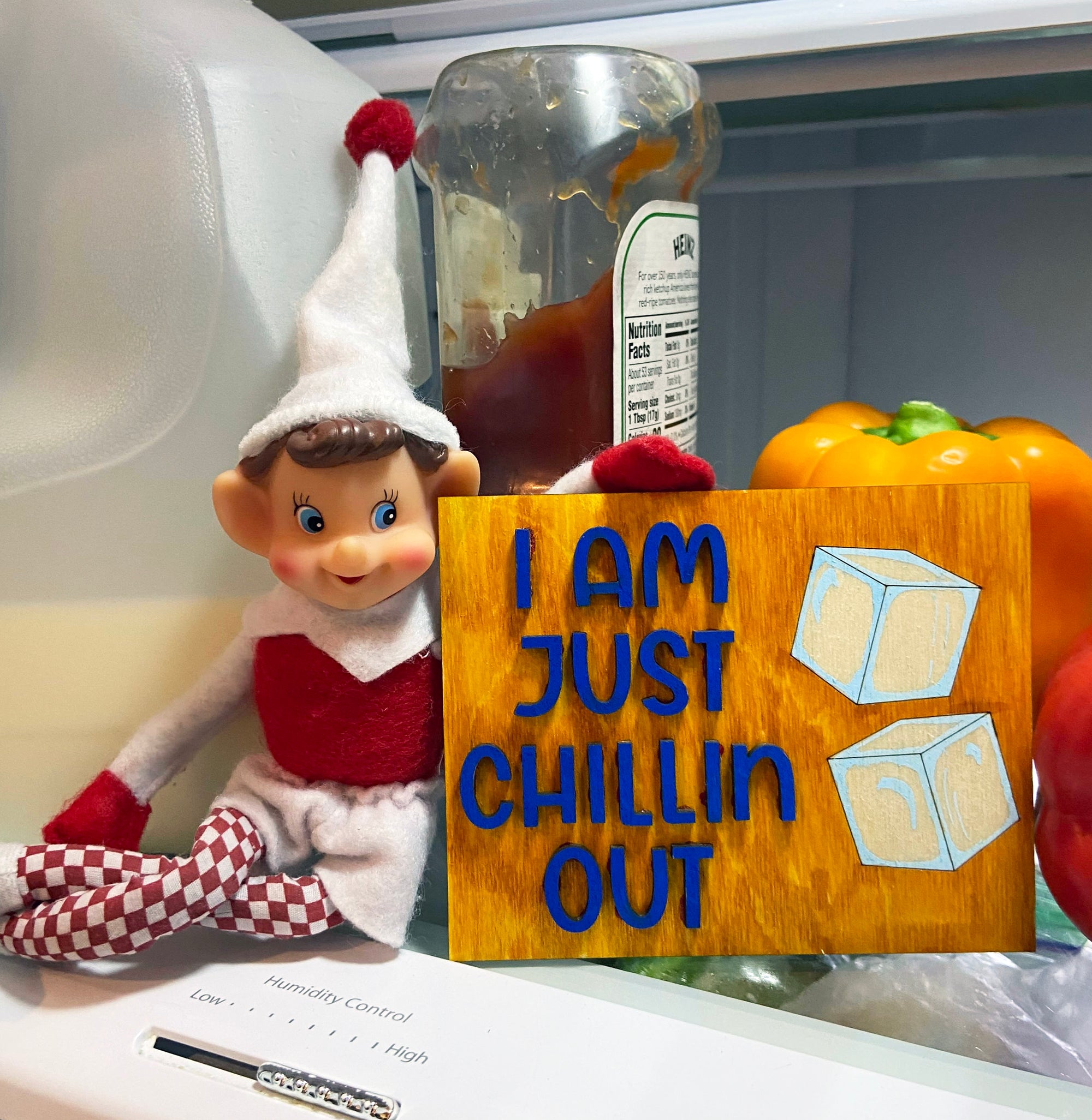 An elf doll from The Rustic Rhode’s DIY Elf Prop Kit sits in a fridge beside veggies, holding an “I AM JUST CHILLIN OUT” sign with ice cubes. A ketchup bottle appears in the background—ideal for festive displays or creative elf scenes.