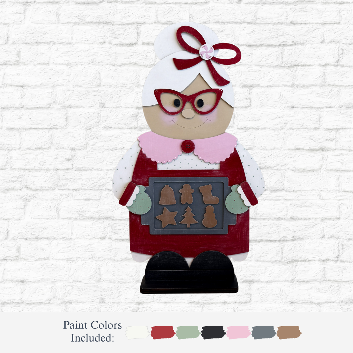 The Rustic Rhode DIY 32 Inch Mrs. Claus features a wooden figure with red glasses and apron, holding a tray of Christmas cookies. Displayed against a white brick wall, it includes labeled paint color swatches below her.