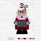 The Rustic Rhode DIY 32 Inch Mrs. Claus features a wooden figure with red glasses and apron, holding a tray of Christmas cookies. Displayed against a white brick wall, it includes labeled paint color swatches below her.