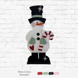 The Rustic Rhode’s DIY 33 Inch Candy Cane Snowman features a black top hat, carrot nose, green mittens, red heart, and festive candy cane—ideal for holiday décor crafts. Included paint colors are shown below the snowman against a white brick wall.