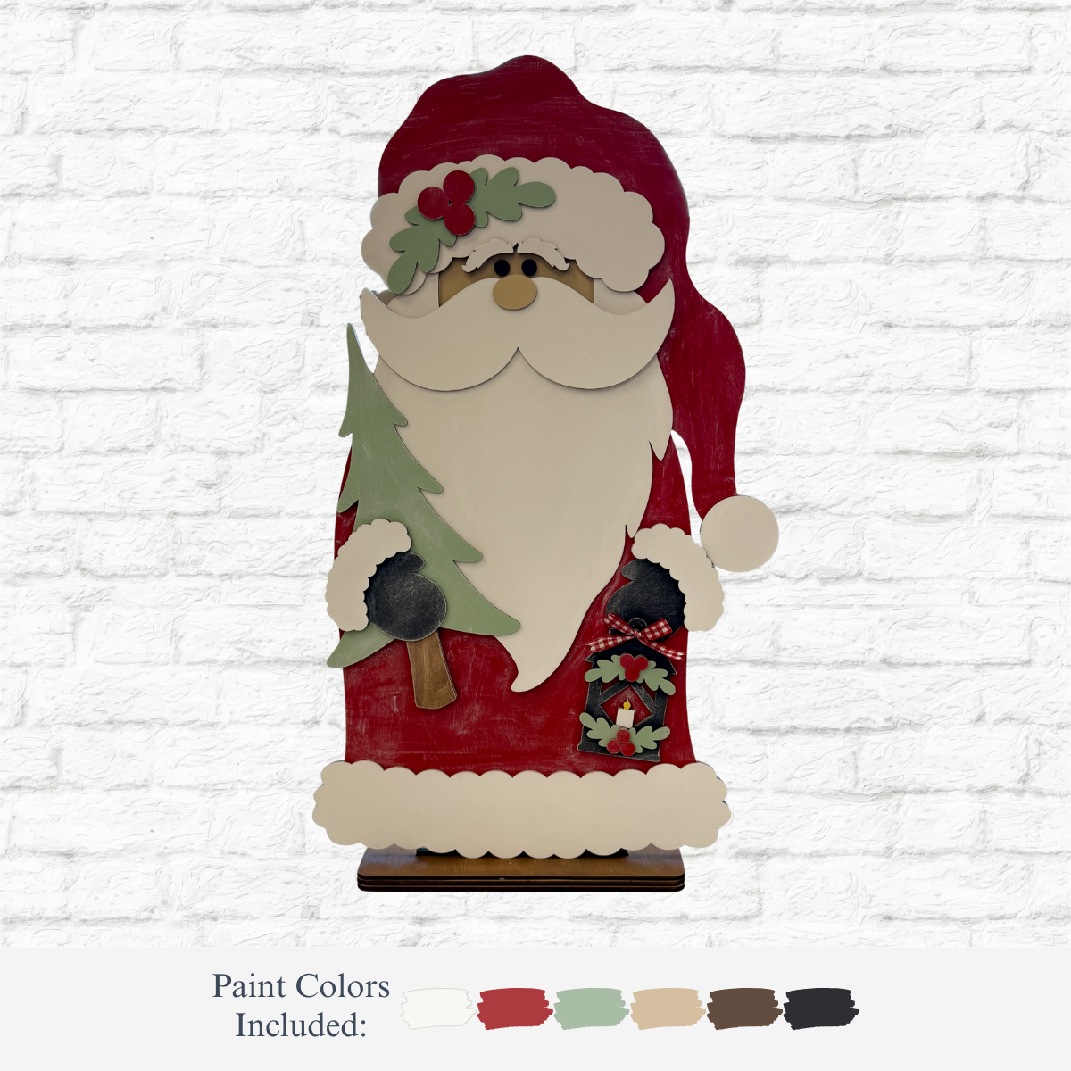 The Rustic Rhode’s DIY 33 Inch Woodland Santa kit features a wooden Santa holding a green tree and black ornament, with holly on his hat, plus paint swatches in red, white, green, beige, brown, and black for customizing your standing figure.