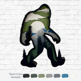 The Rustic Rhode’s DIY 3D Bigfoot Kids Kit includes a Baltic Birch Bigfoot cutout, painted with green trees, gray mountains, a white sun, and clouds, plus six paint colors on a white brick background.