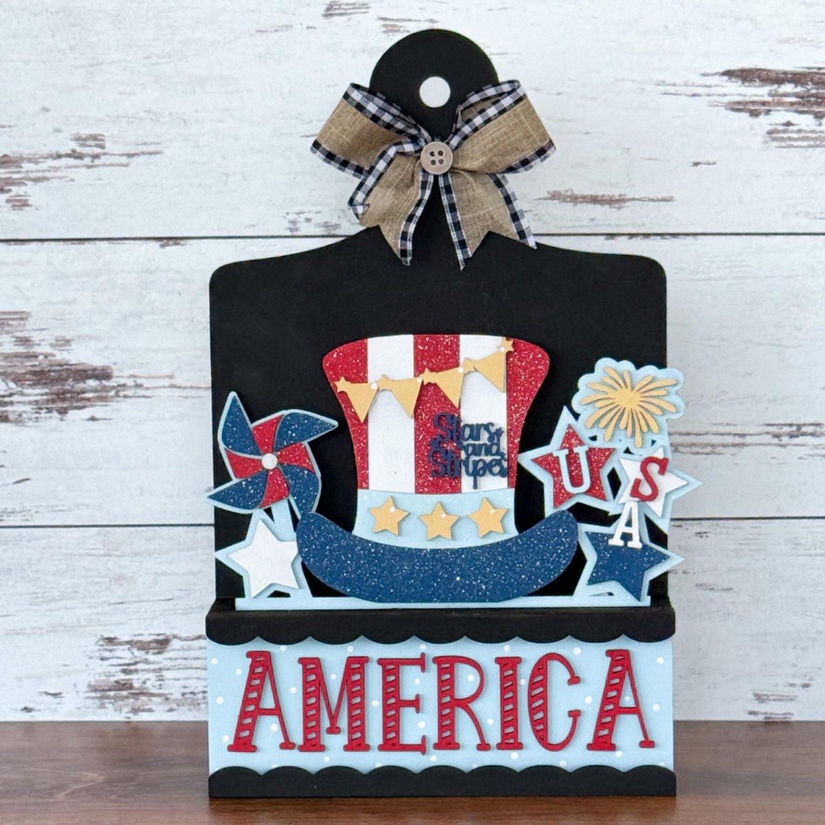 Showcasing The Rustic Rhode’s DIY America 2 Piece Insert, this handmade craft kit features a glittery Uncle Sam hat, fireworks, USA letters, and a plaid bow—perfect for decorating any display against a wood background.