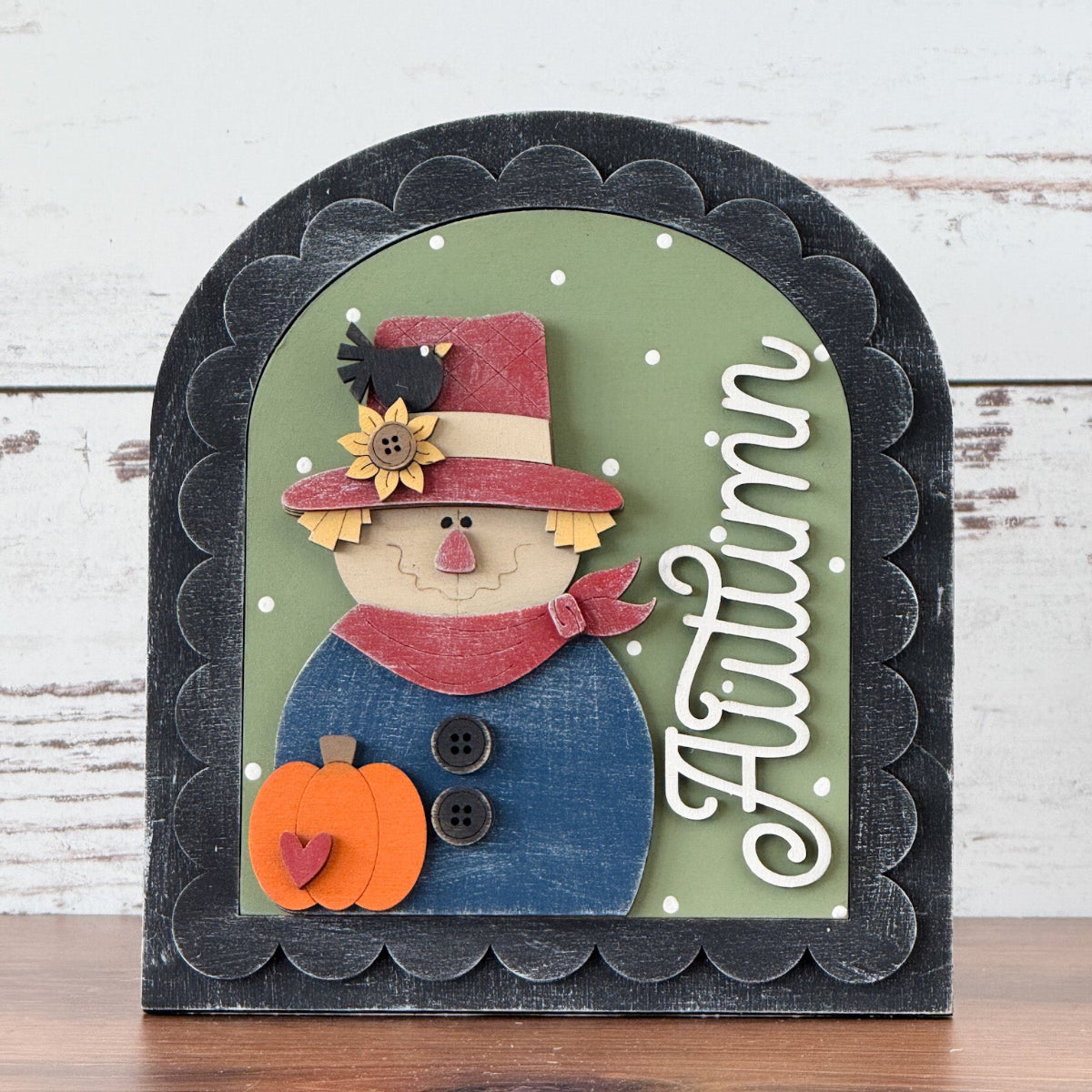 The Rustic Rhode DIY Autumn Scarecrow Arch Insert showcases a smiling scarecrow with a red hat and scarf, a pumpkin, and Autumn in white script on green with white dots, framed by a scalloped black border.