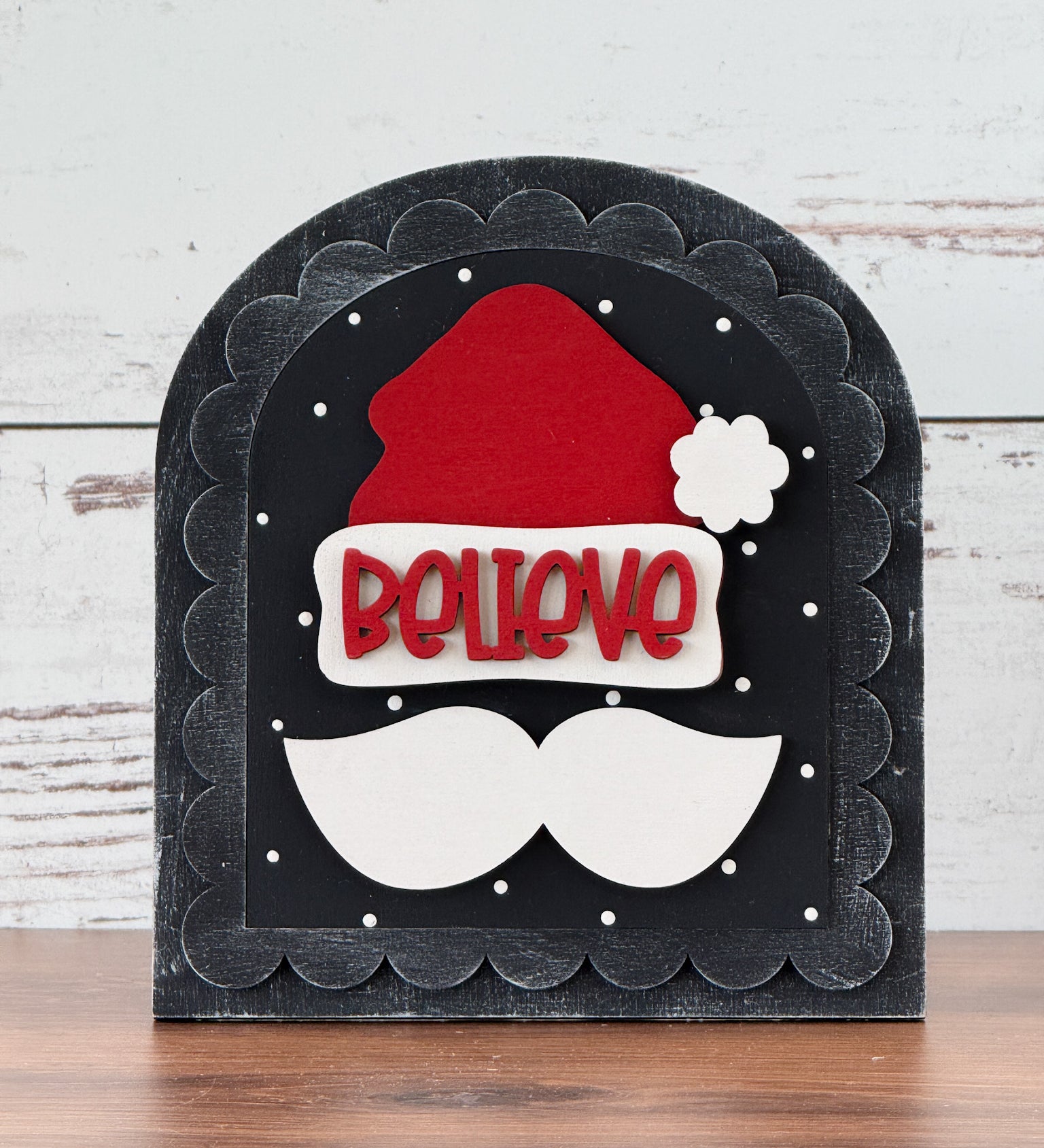The Rustic Rhodes DIY Scalloped Arch Insert Frame features a red Santa hat and white mustache on a black scalloped background with BELIEVE in red letters and white snow-like dots, displayed on a wooden surface.