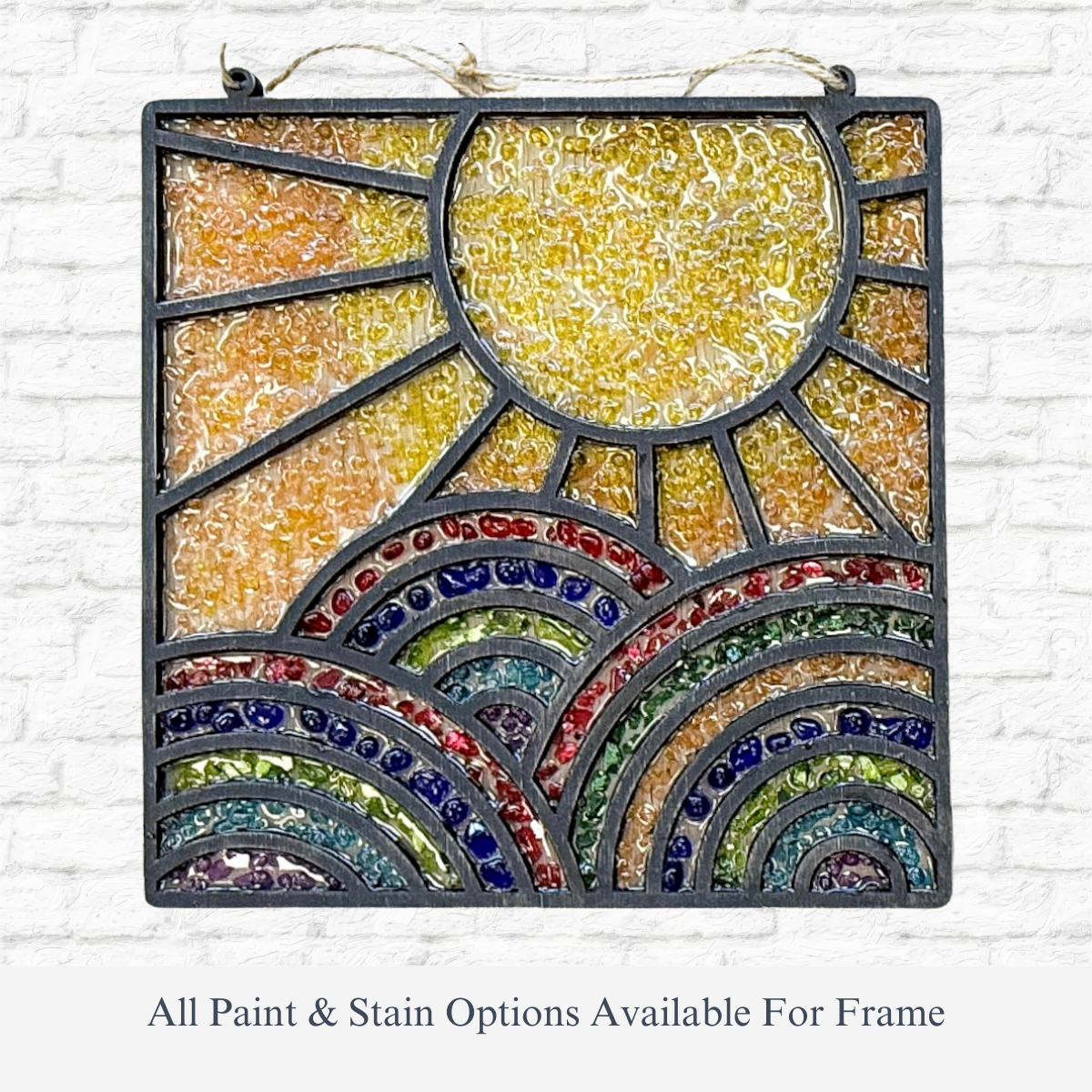 The Rustic Rhodes DIY Chasing Rainbows Faux Suncatcher is a square stained glass art piece with a yellow sun, vibrant arched hills in red, blue, green, and yellow tones, beautifully displayed against a white brick wall.