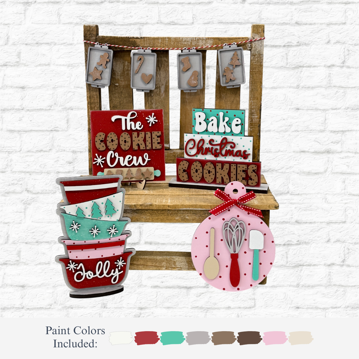 A festive cookie-baking display features The Rustic Rhode’s DIY Christmas Baking Tiered Tray Kit, colorful wooden signs, cookie cutters, garland, and utensils on a rustic chair with paint color samples below on a white brick background.