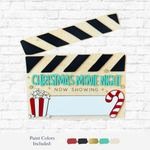 The Rustic Rhode DIY Christmas Movie Night wood clapperboard features “Now Showing” text, popcorn and candy cane graphics—ideal for holiday crafts. Paint color samples are shown below it on a white brick background.
