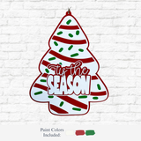 The Rustic Rhode's DIY Christmas Tree Cake Door Hanger, featuring red stripes, green dots, and "Tis the Season" in white and red, hangs on a white brick wall. Red, white, and green paint colors are displayed below.