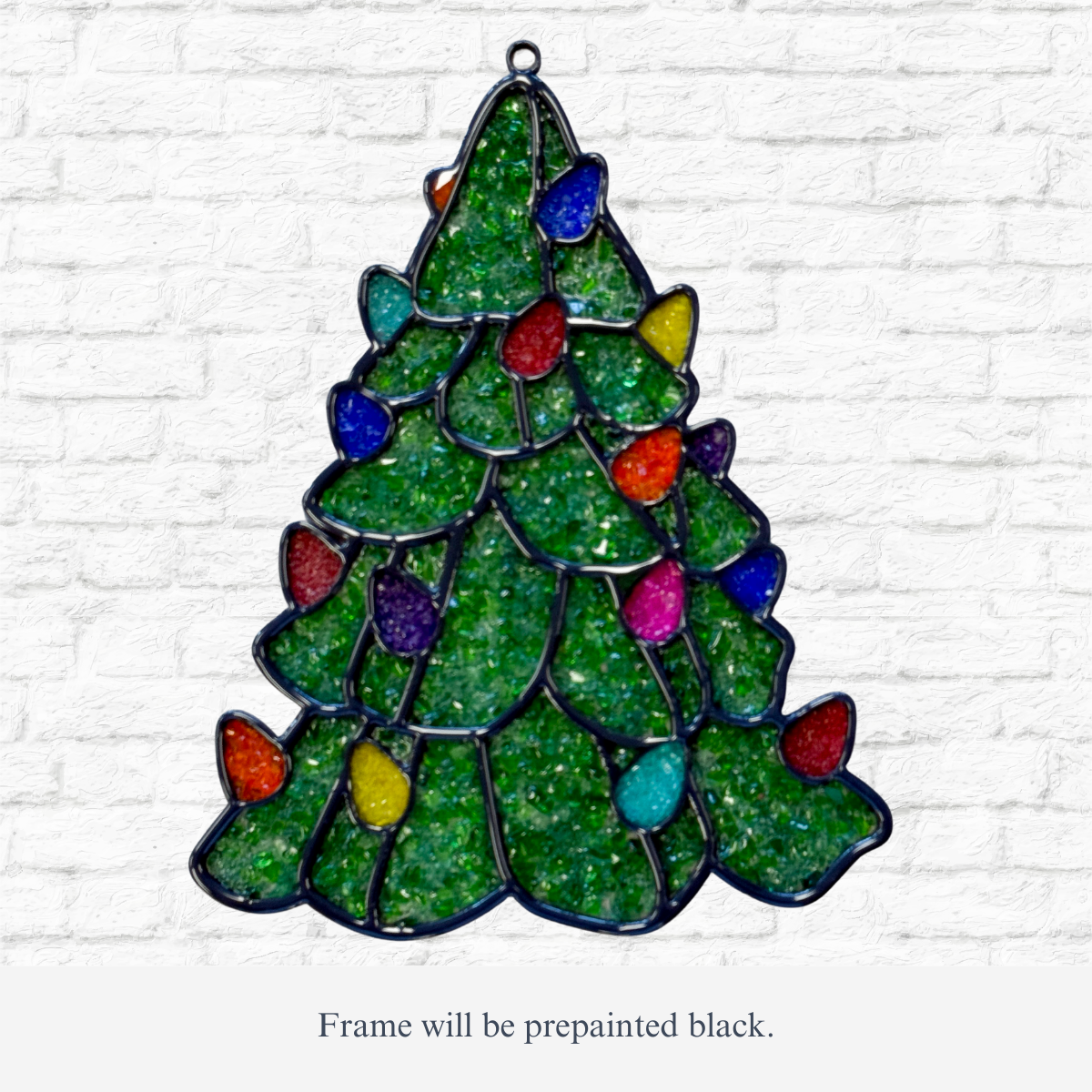 The Rustic Rhode DIY Christmas Tree Faux Suncatcher, shaped like a tree with colorful bulbs and a black metal frame, makes a festive holiday window decoration. Displayed here against a white brick wall.