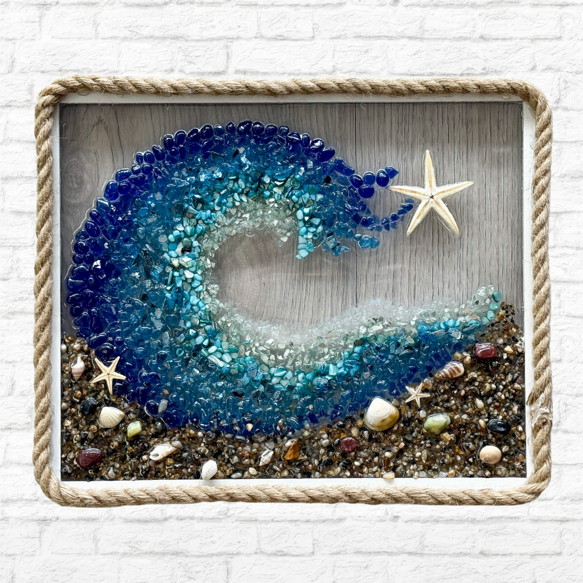 The Rustic Rhode’s DIY Coastal Wave Sea Glass Frame features blue glass waves, a starfish, and seashells on sand. Rope-trimmed framing highlights this handmade coastal art, perfect against a white brick wall backdrop.