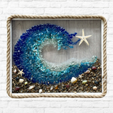 The Rustic Rhode’s DIY Coastal Wave Sea Glass Frame features blue glass waves, a starfish, and seashells on sand. Rope-trimmed framing highlights this handmade coastal art, perfect against a white brick wall backdrop.