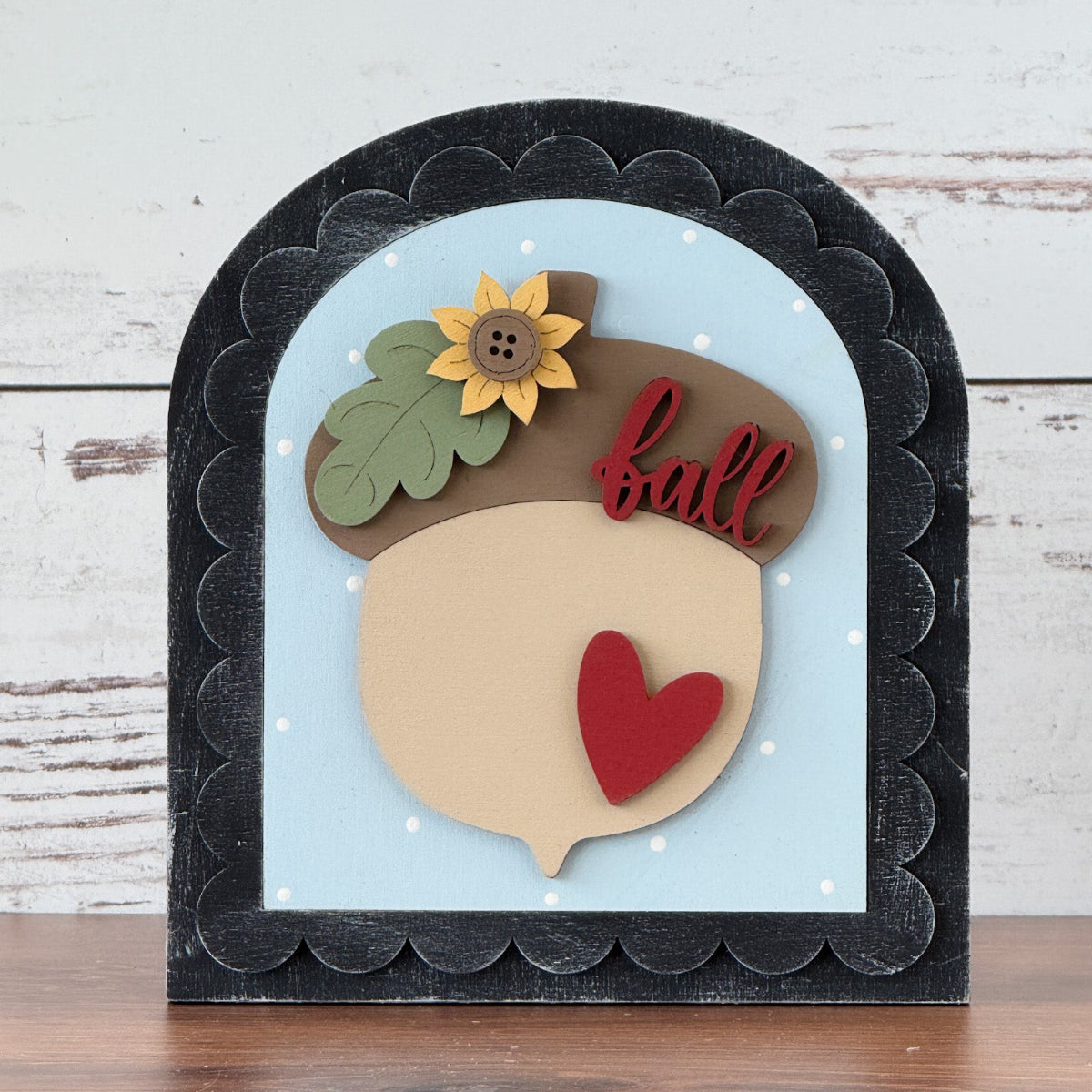 Decorative sign with an acorn design, heart, and sunflower on a wooden surface.