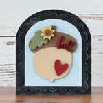 Decorative sign with an acorn design, heart, and sunflower on a wooden surface.