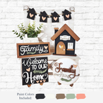 The Rustic Rhode’s DIY Family Home Tiered Tray Kit includes signs saying Family, Welcome to our Home, and Family is everything, plus a heart, leaf motifs, small house, faux plant, and mini house garland on a white brick background.