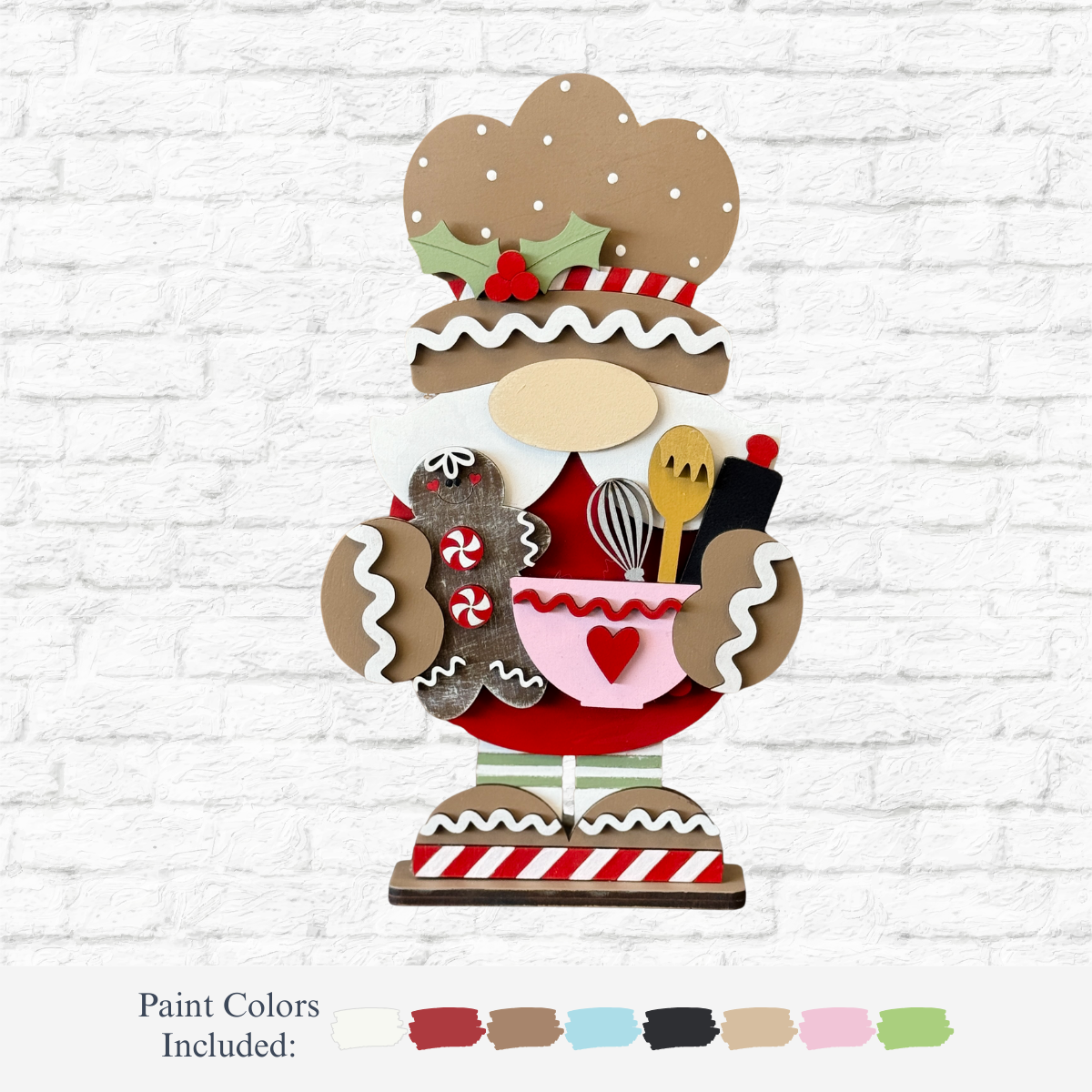 The Rustic Rhode’s DIY Gingerbread Baking Gnome holds kitchen utensils and a mixing bowl, decorated with icing details and peppermint candies. Set against a white brick wall, paint color swatches are displayed below this festive decoration.
