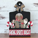 The Rustic Rhode’s DIY Interchangeable Breadboard Frame features a gingerbread figure, candy canes, greenery, and a wooden spoon in a “Baking Spirits Bright” box with a black and burlap bow, set on a white wooden background.
