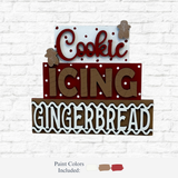 The Rustic Rhode DIY Gingerbread Cookie Word Stacker features “Cookie,” “Icing,” and “Gingerbread” in festive colors with gingerbread men accents, displayed against a white brick wall—ideal for any Holiday Craft Kit display.