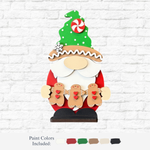 The Rustic Rhode’s DIY Gingerbread Santa Gnome features a wooden gnome with a green hat, white beard, and red coat holding two gingerbread figures—ideal as a festive Christmas decoration.