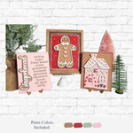 Three gingerbread-themed wooden signs from The Rustic Rhode’s DIY Gingerbread Trio Sign kit—featuring a cookie recipe, a gingerbread person, and a house—stand on easels between two miniature trees in earthy holiday colors.