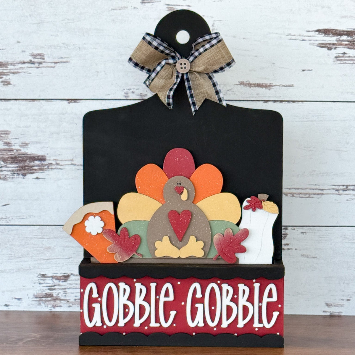 The Rustic Rhode’s DIY Gobble Gobble 2 Piece Insert features a decorative sign with a turkey, pie, leaves, and milk, topped with a plaid bow and “GOBBLE GOBBLE” in white on rustic wood—perfect for handmade or Thanksgiving décor.