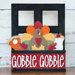 The Rustic Rhode’s DIY Gobble Gobble 2 Piece Insert features a festive turkey with autumn leaves, pie, and drink in a “Gobble Gobble” box—perfect as a Thanksgiving interchangeable window frame decoration.