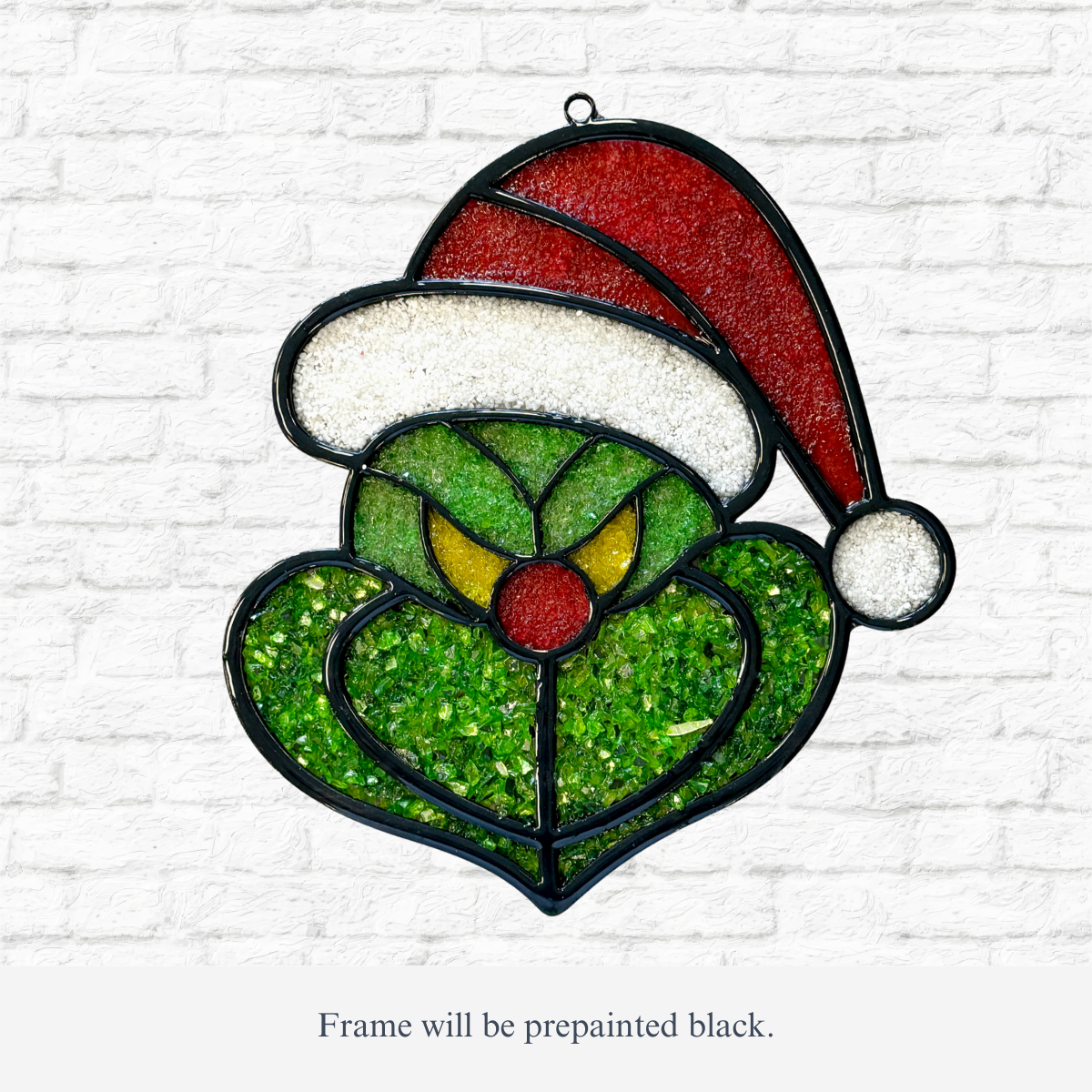 The Rustic Rhode DIY Grinch Faux Suncatcher features a green Grinch with yellow eyes and a red Santa hat, ideal for holiday décor. Shown against a white brick wall. Note: Frame will be prepainted black.