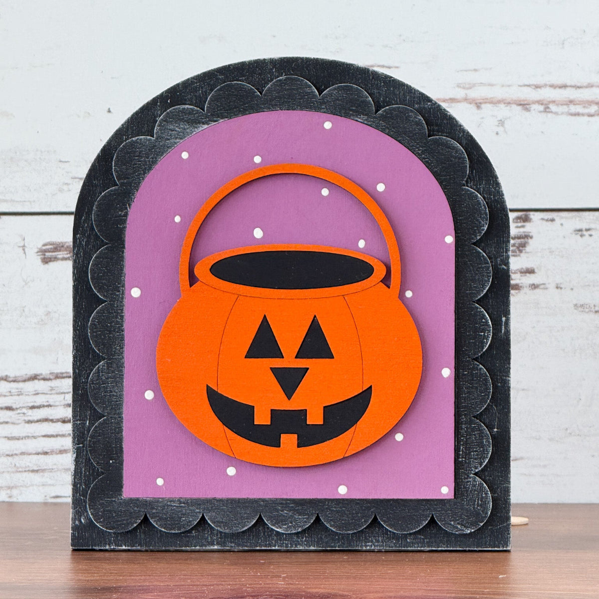 The Rustic Rhode DIY Halloween Jack-O-Lantern Arch Insert features a handcrafted orange jack-o-lantern bucket with a smiling face, set on a purple background with white dots and a scalloped black border atop wood.