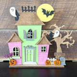 The Rustic Rhode's DIY Haunted House Kit features ghosts, pumpkins, a bat before a yellow moon, a bare tree, a BEWARE sign, and white picket fence accents—all set against a wooden background.