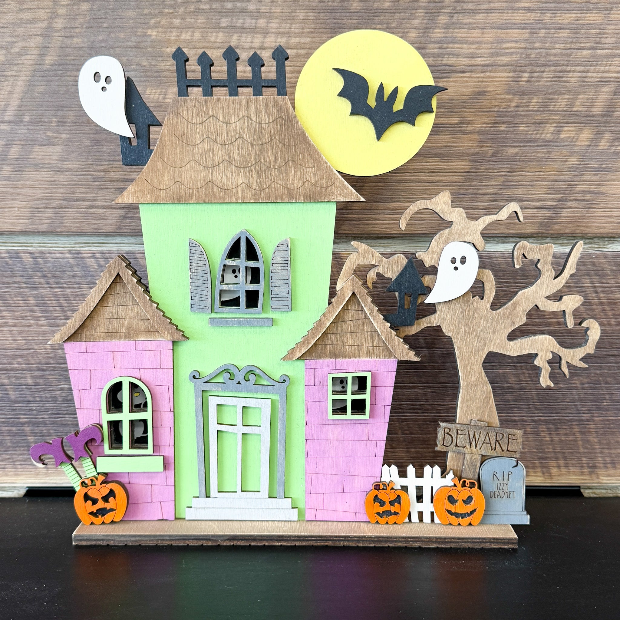 The Rustic Rhode's DIY Haunted House Kit features ghosts, pumpkins, a bat before a yellow moon, a bare tree, a BEWARE sign, and white picket fence accents—all set against a wooden background.