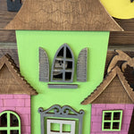 A small ghost peeks from an arched window of a green and pink wooden haunted house with brown roofs. This is the DIY Haunted House Kit by The Rustic Rhode.