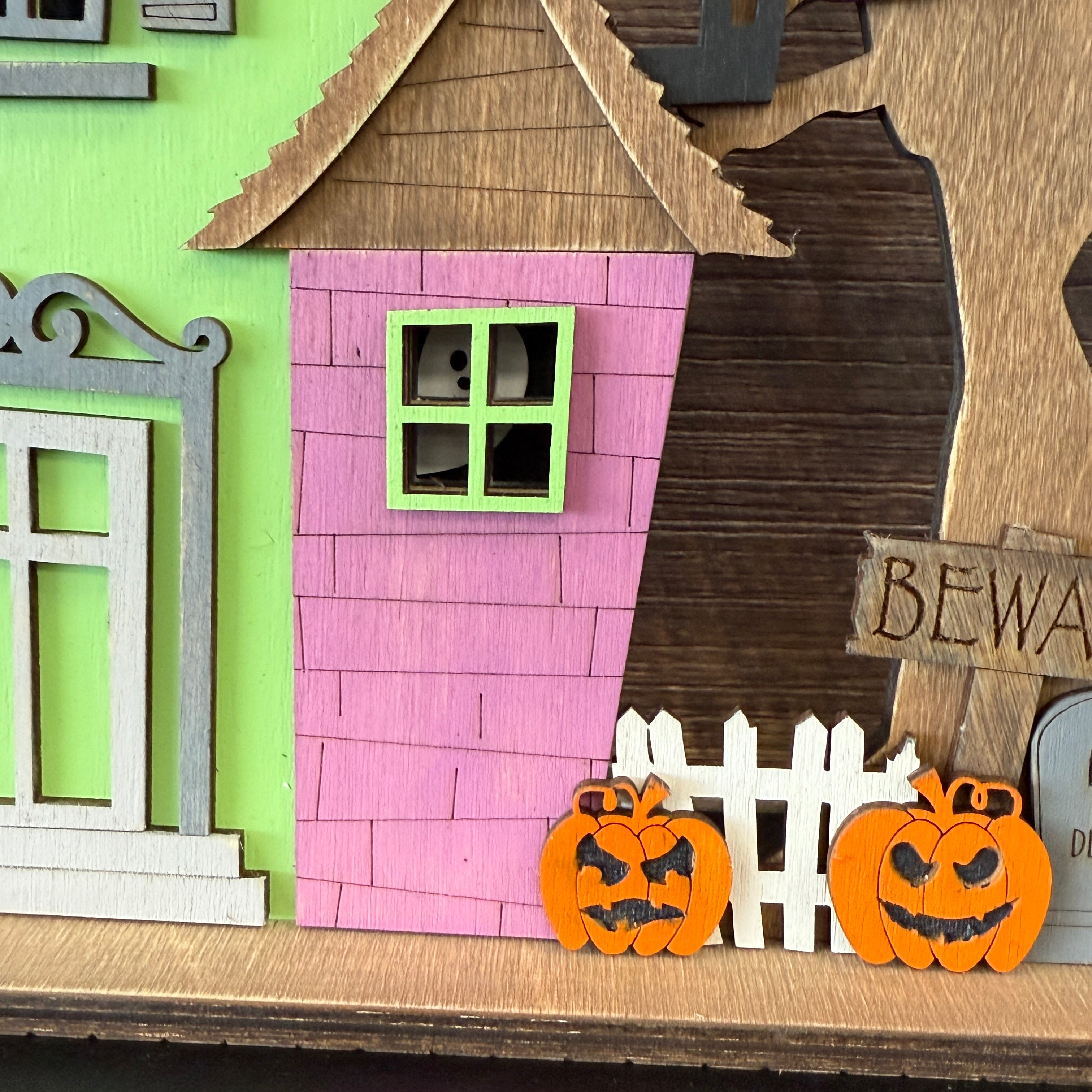 The Rustic Rhode DIY Haunted House Kit features a Halloween wooden decoration with pink and green walls, two carved pumpkins, a white fence, a BEWARE sign, and a ghost peeking from an upstairs window.
