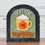 The Rustic Rhode’s DIY Hey There Pumpkin Arch Insert features a mug with whipped cream, a pumpkin design, and playful HEY THERE PUMPKIN text on a green background. This decorative fall sign fits a black scalloped arch insert frame.