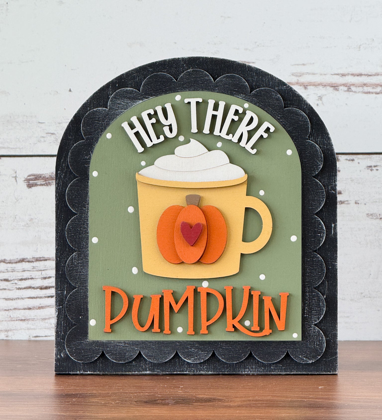 The Rustic Rhode’s DIY Hey There Pumpkin Arch Insert features a mug with whipped cream, a pumpkin design, and playful HEY THERE PUMPKIN text on a green background. This decorative fall sign fits a black scalloped arch insert frame.