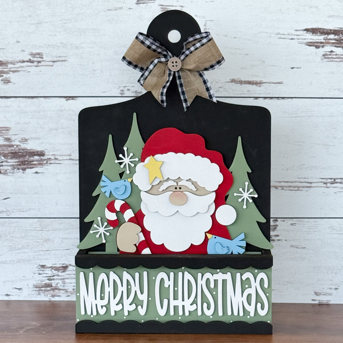 The Rustic Rhode’s DIY Merry Christmas 2 Piece Insert features Santa with a star hat, trees, snowflakes, a plaid bow, and “Merry Christmas”—a festive handmade decoration for your interchangeable window frame.