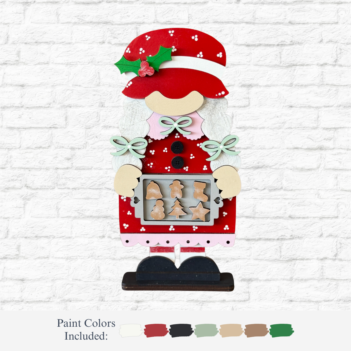 The Rustic Rhode’s DIY Mrs. Claus Baking Gnome is a handmade holiday décor wooden cutout featuring Mrs. Claus with gingerbread cookies in festive red, white, and green; shown here against a white brick wall with paint colors displayed below.